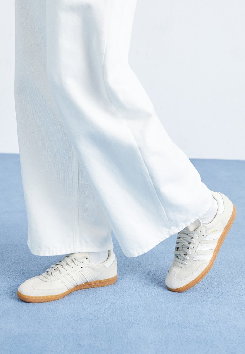 Ljusgrå sneakers med vita ränder och gummisulor, kombinerade med vita vida byxor, stående på en blå matta.