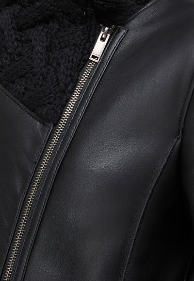 Giacca di pelle nera con zip argento parzialmente aperta, che rivela un spesso tessuto a maglia nera all'interno del colletto.