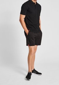 Black polo shirt with short sleeves, paired with black shorts featuring side pockets and red branding. Completed with black athletic shoes.