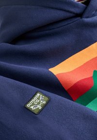 Sudadera azul marino con gráficos de franjas coloridas en naranja, rojo y verde. Presenta un parche rectangular verde con la etiqueta "OUTDOOR CO."
