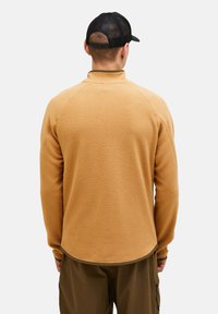 Beiger Fleece-Pullover mit hohem Kragen, Raglanärmeln und strukturiertem Oberflächen, kombiniert mit olivgrünen Hosen. Eine schwarze Mütze vervollständigt den Look.