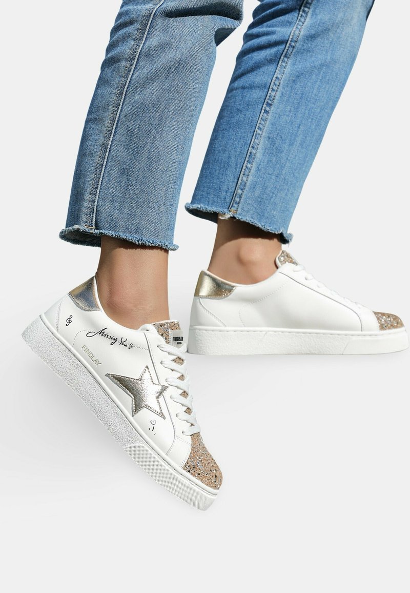 Baskets blanches en cuir avec un accent étoile dorée, un embout beige pailleté et un talon doré métallisé. Présente une semelle texturée blanche et des lacets.