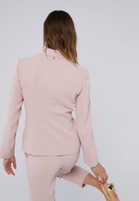 Svijetloružičasti sako krojenog kroja s dugim rukavima, s malim zlatnim hardverskim detaljem straga. Glatka tkanina s utreniranom siluetom.