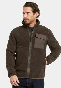 Ruskea fleece-takki, jossa on teksturoitu pinta, täydellinen vetoketju ja rintatasku kontrastiväristä kangasta. Mukana korkea kaulus ja istutetut hihat.