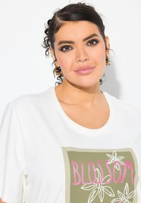 Camiseta blanca con un gráfico rectangular en verde oliva que lleva la palabra "BLOSSOM" en rosa y acentos florales en blanco. Tejido de algodón suave.
