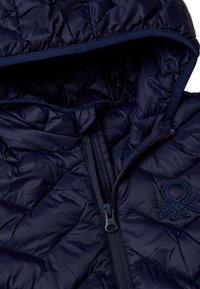 Tmavě modrá prošívaná bunda s kapucí, která má přední zip a vyšívané logo. Materiál má hladkou, lesklou strukturu.