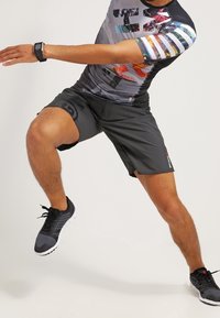 Krátký rukáv sportovního trička s barevným grafickým designem, spárovaný s šedými šortkami až ke kolenům. Model má na sobě černé sportovní boty s bílými podrážkami.