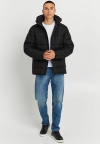 Schwarze Daunenjacke mit Reißverschluss und Kapuze, ausgestattet mit gesteppten Abschnitten, Seitentaschen und hohem Kragen, getragen über einem weißen Hemd.