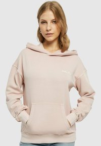Světle růžová mikina s kapucí vyrobená z měkké tkaniny, s přední kapsou, volným střihem a stahovací kapucí. Diskrétní bílé logo na hrudi.