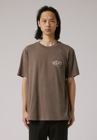 Brown cotton t-shirt with a round neck and short sleeves. Features a small pink graphic on the left chest area. Loose fit and smooth texture.