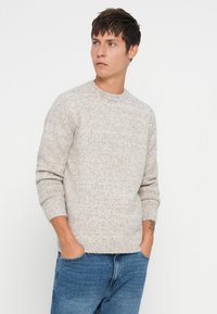 Jeune homme avec des cheveux courts et foncés portant un pull en maille gris clair et un jean bleu, les mains dans les poches, regardant sur le côté devant un fond blanc.