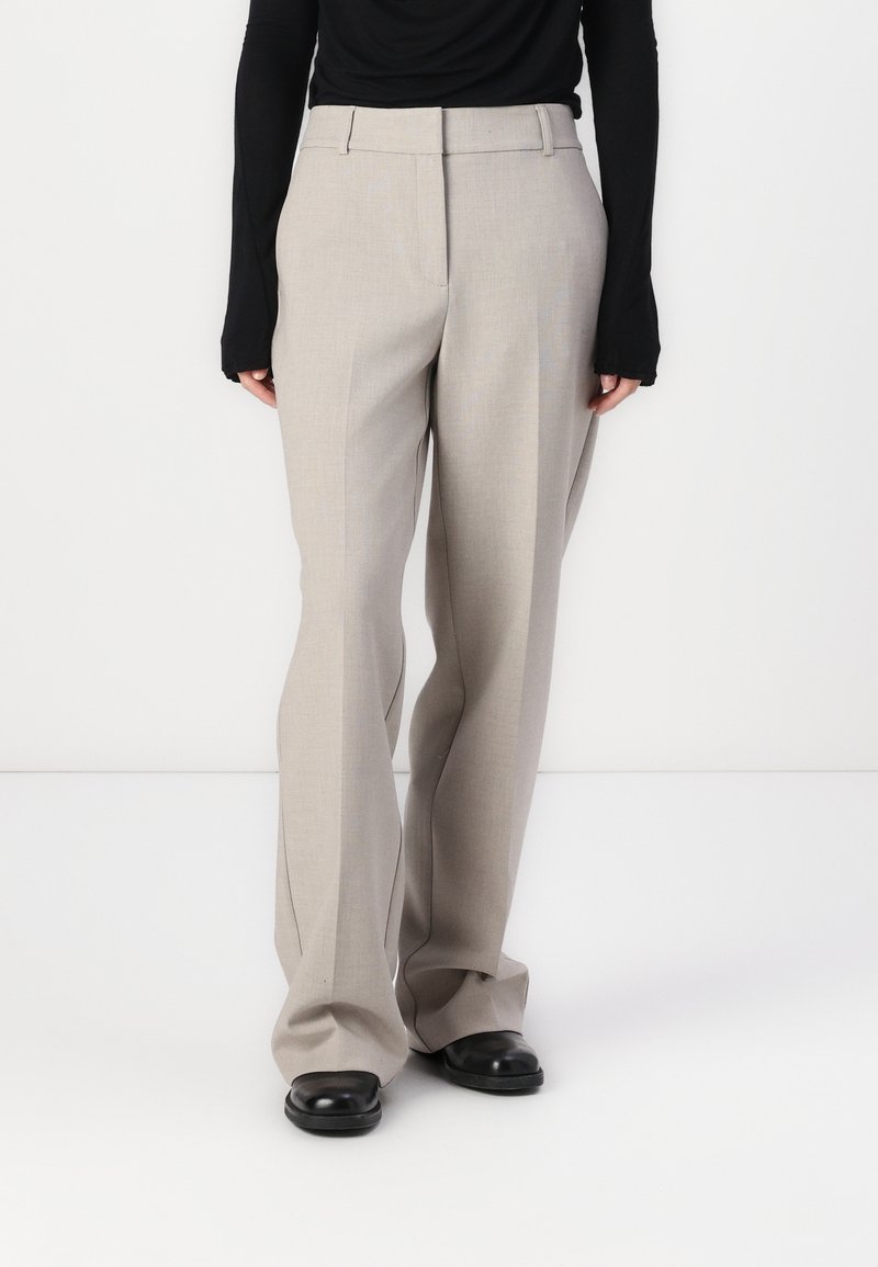 Pantalon taille haute gris clair avec une coupe ajustée, des jambes larges et une texture lisse. Comprend des passants de ceinture et des poches latérales. Porté avec un haut noir.