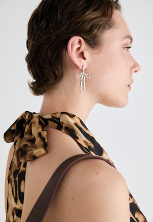 Srebrne naušnice dizajnirane s izduženim oblicima i detaljem mašne, prikazane na pozadini bluze s leopard printom.