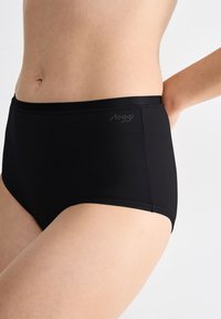 Gros plan sur le bas du torse d'une personne portant un sous-vêtement noir taille haute avec un petit logo "sloggi" gris sur le côté gauche.