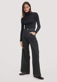 Zwarte coltrui gecombineerd met hooggepuntte, wijde donkere denim jeans. Model staat met één hand in de zak en donkere schoenen. Lichte achtergrond.