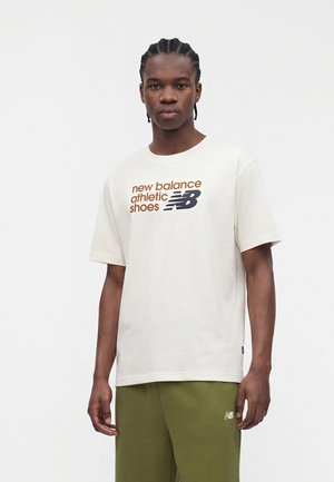 Jeune homme aux cheveux tressés portant un t-shirt graphique blanc New Balance et un pantalon vert olive sur un fond blanc uni.