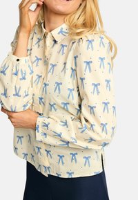 Kremna bluza z vzorcem ponavljajočih se modrih trakov, ovratnikom na gumbe, nabranimi rokavi in rahlo ukrivljenim spodnjim robom. Izdelana je iz lahkega blaga.