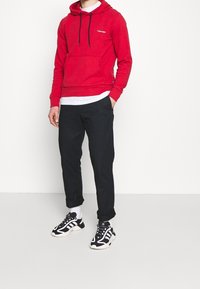 Sudadera roja con capucha, bolsillo frontal y cordones, combinada con pantalones negros y zapatillas deportivas negras con detalles y cordones blancos.