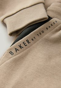 Beżowa tkanina z czarną podszewką oraz napisem "BAKER BY TED BAKER" nadrukowanym na beżowym pasku blisko mankietu odzieży.