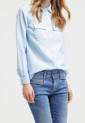 Kobieta ubrana w jasnoniebieską koszulę zapinaną na guziki z kieszeniami na piersi i niebieskie dżinsy, stojąca na jednolitym białym tle.