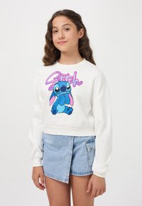 Felpa bianca corta con un personaggio dei cartoni animati blu in posizione seduta, con accenti rosa e la parola "Stitch" in grassetto rosa.