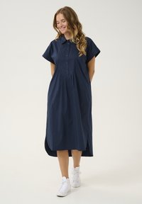 Rochie bleumarin, cu nasturi, mâneci scurte, guler și fante laterale. Țesătura pare ușoară și are o textură netedă. Se asortează cu adidași albi.