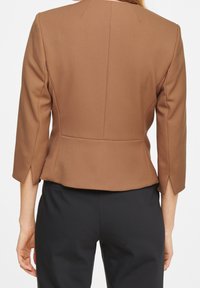 Femme portant un blazer marron ajusté à manches trois-quarts avec des fentes latérales, associé à un pantalon noir, vue de dos.