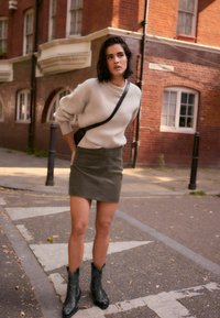 Knit cream sweater with ribbed texture, olive green mini skirt, and black cowboy boots with stitching details, set against a brick building.