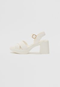 Sandale blanche à talon bloc, avec deux brides croisées et une bride de cheville avec une boucle dorée. Matériau lisse et design minimal.