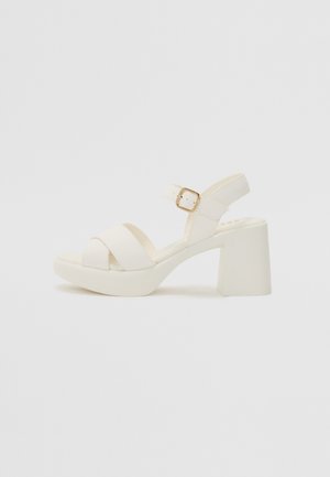 White heeled sandal with a block heel, featuring two crossed straps and an ankle strap with a gold buckle. Smooth material and minimal design.