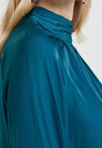 Blouse teal avec un col haut à volants et un tissu fluide. Le matériau a une texture lisse et tombe doucement sur l'épaule.