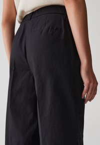 Pantalons larges noirs avec une finition texturée, présentant une poche arrière et une taille douce, montrés de côté avec une main reposant sur la cuisse.