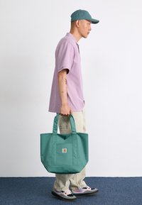Teal lærred tote taske med to håndtag, der har en frontlomme og et logo mærke. Tasken holdes af en person iført lyse bukser og sneakers.