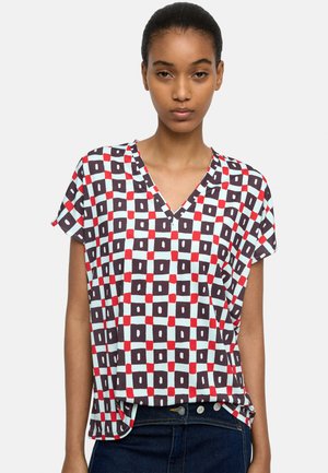 Młoda kobieta w bluzce z krótkim rękawem i dekoltem w serek, z geometrycznym wzorem kwadratów w kolorach czerwonym, czarnym i białym, w parze z ciemnoniebieskimi dżinsami.