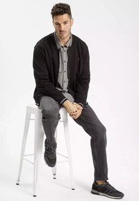 Czarny kardigan nałożony na koszulę w kratę, szare dżinsy i trampki; siedzący na białym stołku, neutralne tło, swobodny strój.