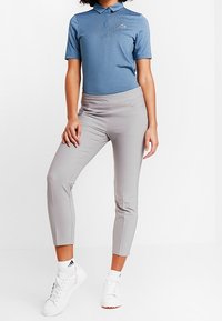 Person som bär en blårandig polo-tröja, ljusgrå välsittande capribyxor, vita Adidas-strumpor och vita sneakers, stående mot en vit bakgrund.