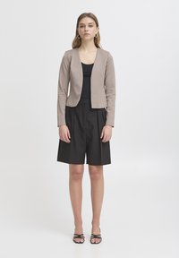 Femme debout, portant un blazer taupe, un haut noir, un short plissé noir mi-genoux, des boucles d'oreilles créoles et des talons noirs à brides, avec un fond neutre.