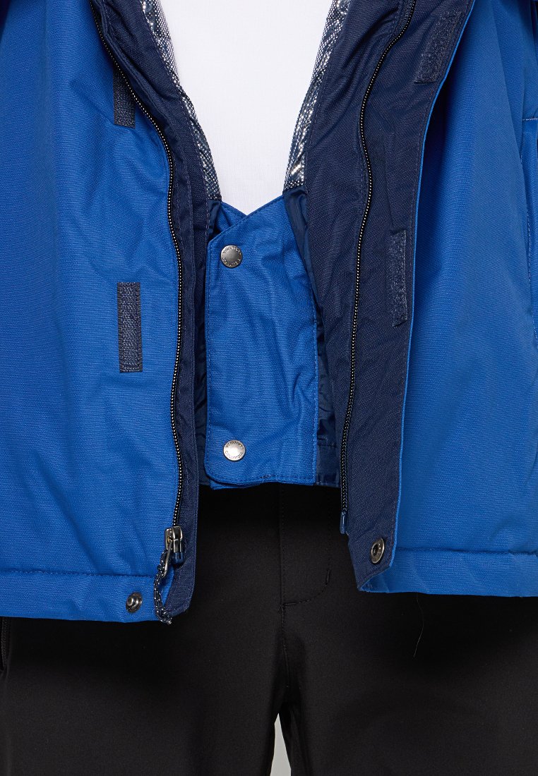 Blauwe jas met binnenflap die is vastgemaakt met twee metalen drukknoopjes, gedeeltelijk open geritst over zwarte broeken en een witte blouse.