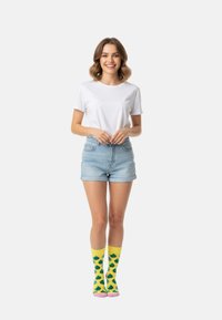 Witte t-shirt, blauwe denim short, en gele sokken met groene patronen. Model staat met de handen gevouwen, wat de details van de outfit benadrukt.