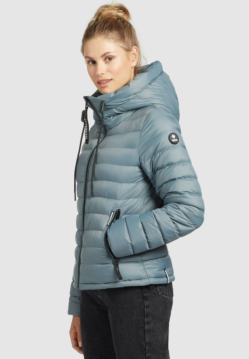 Frau mit blonden Haaren im Dutt, die eine hellblaue gesteppte Jacke mit hohem Kragen und schwarze Jeans trägt, steht vor einem hellen Hintergrund.