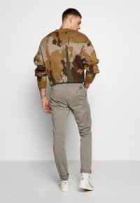 Rjav in zeleno vzorčast pleten pulover, s sproščenim krojem, v kombinaciji z svetlo sivo slim-fit chinos in belimi športniki.