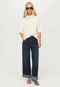 Top bianco a maniche corte con collo alto, abbinato a jeans ampi blu scuro con risvolti e sandali neri con cinturini.