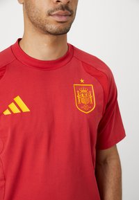 Κόκκινο αθλητικό μπλουζάκι με στρογγυλή λαιμόκοψη, με το κίτρινο λογότυπο της Adidas και το κίτρινο έμβλημα της ισπανικής εθνικής ομά�δας με στέμμα.