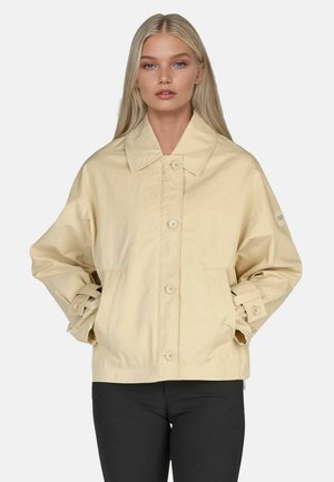 Blonde woman wearing beige button-up jacket with collar and pockets, paired with black pants, standing with hands in pockets.