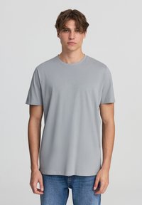 Szara koszulka z krótkim rękawem, wykonana z miękkiego materiału, z okrągłym dekoltem i luźnym krojem. Noszona z niebieskimi dżinsami.