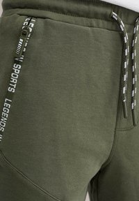 Olivové jogger nohavice s textúrovanou tkaninou, bočná zipsová vrecko a šnúrka so bielymi a čiernymi akcentmi. Obsahuje branding pozdĺž vrecka.