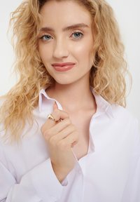 White button-up shirt with a relaxed fit, featuring a folded collar. The model wears a gold ring, finger resting on the shirt's collar.