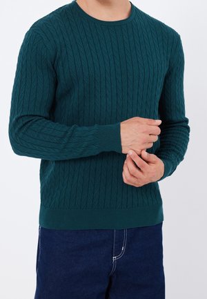 Homme portant un pull torsadé bleu sarcelle avec poignets et ourlet côtelés, ajustant la manche, associé à un jean bleu foncé.
