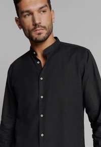 Zwarte button-up blouse met een staande kraag, gemaakt van een lichte stof. Heeft een subtiele textuur en een klein merkaccent op de borst.