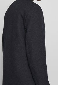 Mørk marineblå sweater med struktureret strik, der har en rund halsudskæring og tætsiddende ærmer. Stoffet ser blødt og tykt ud.